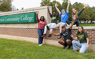 Six diverse students pose excitedly with their hands up near a brick wall with a sign partially visible that reads "erick Community College," likely Frederick Community College. Three students are sitting on the wall, and three are kneeling or crouching in front of it in the grass.